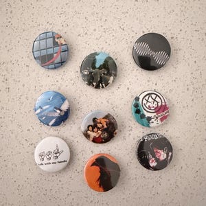May include: A collection of nine circular pin-back buttons featuring various graphic designs, including a grid pattern, a landscape, a sound wave, a bird, a group of people, a smiley face, sign language hands with the text talk with my hands.
