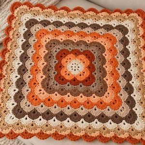 May include: A square crochet blanket featuring a concentric pattern of scalloped edges in shades of burnt orange, dark brown, tan, and cream. The design radiates from a central flower motif, with each layer expanding outward in a repeating shell stitch.