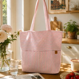 May include: A rectangular tote bag featuring a pattern of thin red horizontal stripes on the upper section and vertical red stripes on the lower section. The bag includes two long shoulder straps and a small brown rectangular label on the front.