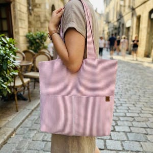 May include: A large tote bag featuring a red and white pinstripe pattern. The bag has two shoulder straps and a front pocket with a small brown rectangular label attached to the right side. The fabric displays horizontal stripes on top and vertical stripes below.