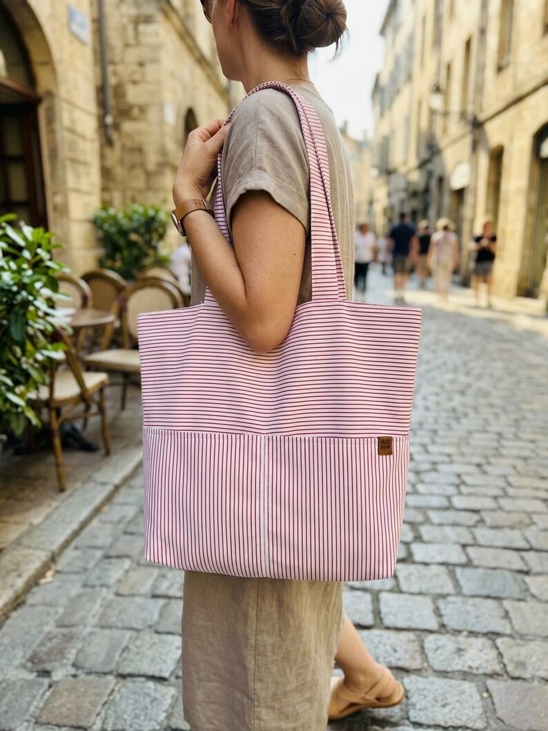 May include: A large tote bag featuring a red and white pinstripe pattern. The bag has two shoulder straps and a front pocket with a small brown rectangular label attached to the right side. The fabric displays horizontal stripes on top and vertical stripes below.