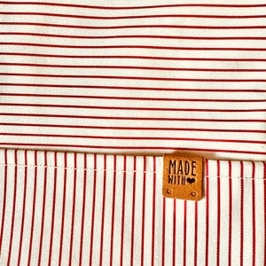 May include: A fabric textile featuring a pattern of thin red horizontal and vertical stripes. A small, rectangular tan leather-like tag is stitched onto the fabric, embossed with the text "MADE WITH" followed by a small heart symbol.