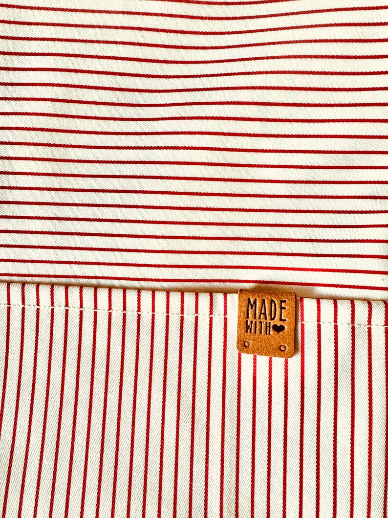 May include: A fabric textile featuring a pattern of thin red horizontal and vertical stripes. A small, rectangular tan leather-like tag is stitched onto the fabric, embossed with the text "MADE WITH" followed by a small heart symbol.