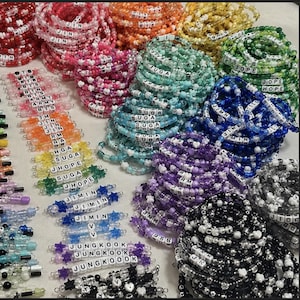 May include: A collection of colorful beaded bracelets featuring white letter beads that spell out names like JIN, SUGA, JHOPE, JIMIN, V, and JUNGKOOK. The bracelets are made with translucent, multi-colored plastic beads and star-shaped accents.