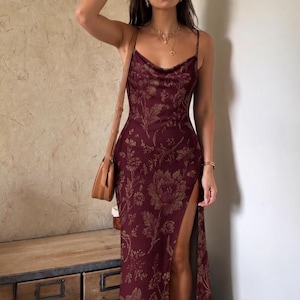 May include: A floor-length burgundy slip dress featuring a tonal floral pattern, a cowl neckline, thin spaghetti straps, and a high side slit. The garment is paired with a tan leather shoulder bag, gold layered necklaces, and gold bracelets.