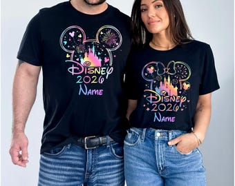 May include: Two black t-shirts featuring a colorful, gradient-style graphic of a castle silhouette inside a Mickey Mouse ear outline. The design includes fireworks, stars, and the text Disney 2026 Name in a vibrant, multi-colored font style.