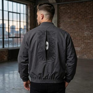 Puede incluir: Una chaqueta bomber gris carbón con una abertura vertical en el panel trasero que revela una inserción de tela blanca estampada con un gráfico negro de un ojo rodeado de símbolos circulares, rayos y patrones de plumas estilizados.