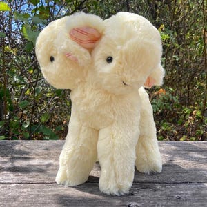 May include: A plush toy featuring two heads on a single body, crafted from cream-coloured, soft, fuzzy material. The creature has small black eyes and inner ears accented with a subtle pink and white striped pattern, standing on four legs.