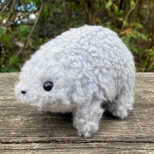 May include: A small, light grey plush toy shaped like a rounded animal with a fuzzy, textured exterior. It features a single visible dark, round eye and a tiny, dark nose. The creature has a compact, stout body with short, stubby legs.