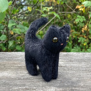 May include: A small, handcrafted black cat figurine with a fuzzy, textured surface. The cat has a raised tail, pointed ears, and two large, round, amber-colored glass eyes. A tiny pink dot is visible on the face, representing a small nose.