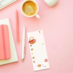 May include: A rectangular notepad with horizontal lines, featuring a picnic basket, strawberries, a bee, and bear faces. The bottom of the notepad displays the text 'B rry Sweet' with a small bear illustration. The notepad is white with colourful graphics.