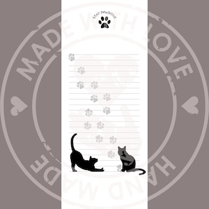 May include: A white rectangular notepad with horizontal lines, featuring a black paw print at the top with the text STAY PAWSITIVE. The page includes a light gray heart outline, scattered gray paw prints, and two black cat silhouettes at the bottom.