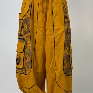 May include: Mustard yellow cargo trousers featuring a drawstring elastic waistband and large side pockets with black piping. The side panels display a brown and black geometric pattern, complemented by additional zippered and flap-covered pockets.