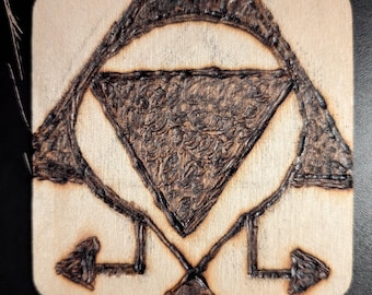 Può includere: Un sottobicchiere quadrato in legno con un design scuro pirografato che raffigura un triangolo centrale racchiuso in un arco circolare, con due piccoli simboli a forma di freccia rivolti verso l'esterno negli angoli inferiori.