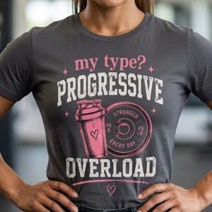 May include: A charcoal grey short-sleeve t-shirt featuring a graphic print with the text "my type? PROGRESSIVE OVERLOAD". The design includes a pink shaker bottle and a weight plate with the text "STRONGER EVERY DAY" and "45 LB".