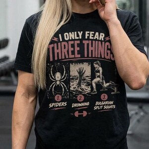 Pode incluir: Uma t-shirt preta de manga curta com o texto I ONLY FEAR THREE THINGS em rosa. Abaixo, três ilustrações: uma aranha, uma mão a sair da água e uma pessoa a fazer agachamentos búlgaros, numeradas de 1 a 3.
