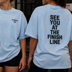 May include: A light blue short-sleeve t-shirt featuring the black text HYROX EST 2022 on the front left chest and the black text SEE YOU AT THE FINISH LINE printed in a large vertical block on the back of the garment.