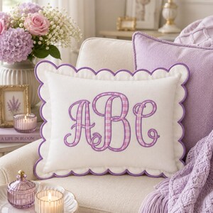 May include: A rectangular white throw pillow featuring a scalloped edge trimmed in purple. The center displays a large, embroidered monogram of the letters ABC in a purple and white gingham pattern with a decorative, swirling script font.