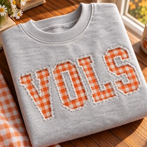 May include: A heather gray crewneck sweatshirt featuring the word VOLS in large, block-style letters. The letters are filled with an orange and white gingham pattern and are outlined with thick, white decorative stitching.