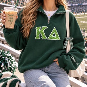 May include: A dark green quarter-zip sweatshirt featuring the Greek letters KA in white with a green polka dot pattern. The left sleeve is embroidered with the text Theta Pi in white. A white tote bag with a green and white gingham bow is present.