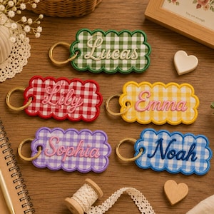 May include: Five rectangular keychains with scalloped edges and a gingham pattern in green, red, yellow, purple, and blue. Each keychain features a gold-tone metal ring and is embroidered with a name: Lucas, Lily, Emma, Sophia, or Noah.