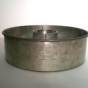 May include: A round, metallic silver-toned cake pan with a central tube, featuring a weathered finish and the embossed text Cream City on the side. The baking pan has a wide, circular rim and a hollow center column for even heat distribution.
