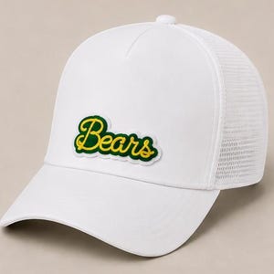 May include: A white trucker hat featuring a mesh back panel and a solid front panel. An embroidered patch is centered on the front, displaying the word Bears in yellow cursive script with a dark green outline and a white border.