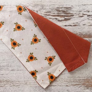May include: A triangular pet bandana featuring a cream-colored fabric side with a repeating pattern of orange sunflowers and small brown dots, and a solid burnt orange fabric on the reverse side, showing a stitched seam along the folded edge.