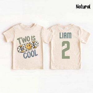 May include: A natural-colored t-shirt featuring a graphic of a checkered skateboard with a yellow smiley face and the text Two is Cool on the front, and the name Liam with the number 2 in green on the back. The word Natural is printed at the top.