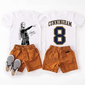 May include: Two white t-shirts and two pairs of brown shorts with a pair of black and white checkered slip-on shoes. One shirt features a black graphic of a basketball player with the number 8 and a signature. The other shirt displays CUNNINGHAM 8.
