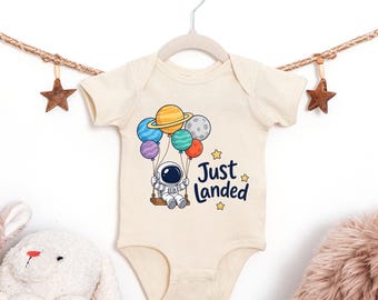 Puede incluir: Un body de bebé de manga corta en color natural con un gráfico de un astronauta sentado en un columpio sostenido por globos de colores con forma de planetas. El texto Just Landed aparece en azul oscuro junto al astronauta y pequeñas estrellas.