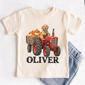 May include: A cream-coloured short-sleeve t-shirt featuring a graphic of a golden retriever puppy wearing a green checkered bandana, sitting on a red tractor pulling a wooden trailer filled with orange pumpkins, with the name OLIVER printed below.