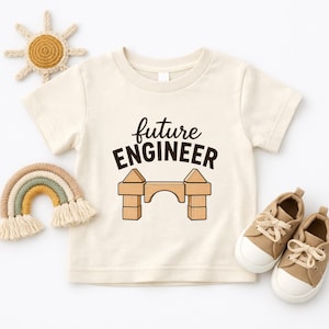 May include: A cream-coloured short-sleeve t-shirt featuring the text "future ENGINEER" in dark brown letters above a graphic of a bridge constructed from light brown wooden building blocks. The shirt is displayed with a crocheted sun and rainbow.