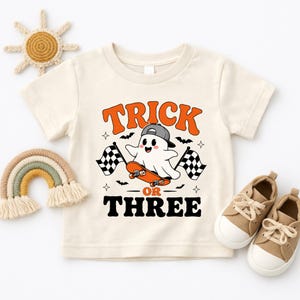 May include: A cream-colored short-sleeve t-shirt featuring a graphic of a white ghost wearing a gray baseball cap while riding an orange skateboard. The text reads TRICK OR THREE in orange and black, flanked by two black and white checkered flags.