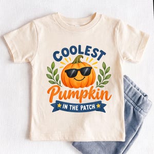 May include: A cream-colored short-sleeve t-shirt featuring a graphic of an orange pumpkin wearing black sunglasses. The text reads "COOLEST PUMPKIN IN THE PATCH" in blue and orange lettering, accompanied by green leaf illustrations and yellow stars.
