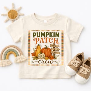 May include: A cream-colored short-sleeve t-shirt featuring a graphic print of pumpkins, autumn leaves, and a wooden signpost with the text "FUN FALL MEMORIES". The design includes the words "PUMPKIN PATCH crew" in orange and green lettering.