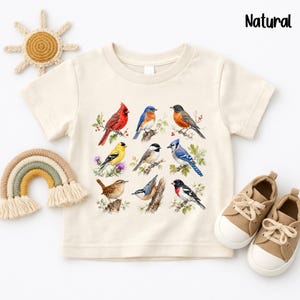 May include: A natural-colored short-sleeve t-shirt featuring a graphic print of nine different bird species perched on branches. The birds include a red cardinal, bluebird, robin, goldfinch, chickadee, blue jay, wren, nuthatch, and grosbeak.