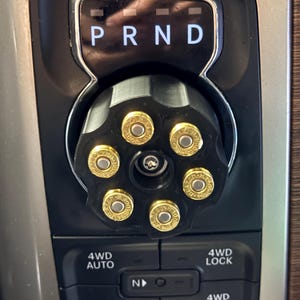 May include: A black vehicle gear shift knob shaped like a revolver cylinder with six gold-colored bullet casings inserted. The knob is positioned below a gear indicator panel displaying P R N D and above a 4WD control panel with 4WD AUTO, 4WD LOCK, 2WD, and 4WD LOW settings.