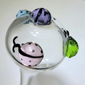May include: A clear glass ornament with a pink, purple, blue, and green ladybug design. The ornament is shaped like a sphere with a long stem.
