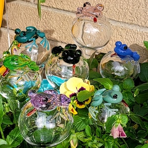 May include: A collection of six clear glass plant watering globes, each topped with a colorful glass frog figurine. The frogs are colored in shades of teal, light pink, bright green, black, blue, and pale green, each featuring a small red tongue.