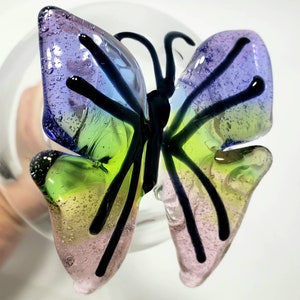 May include: A glass butterfly ornament with iridescent wings in shades of purple, blue, green, and pink. The butterfly has black veins and is attached to a clear glass sphere.
