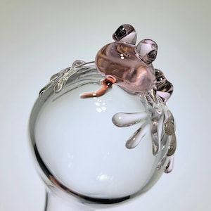 May include: A clear glass sphere featuring a sculpted, light pink glass frog perched on the top edge. The frog has two prominent, rounded eyes and a small, curved tail extending downward. The glass is smooth, transparent, and reflective.