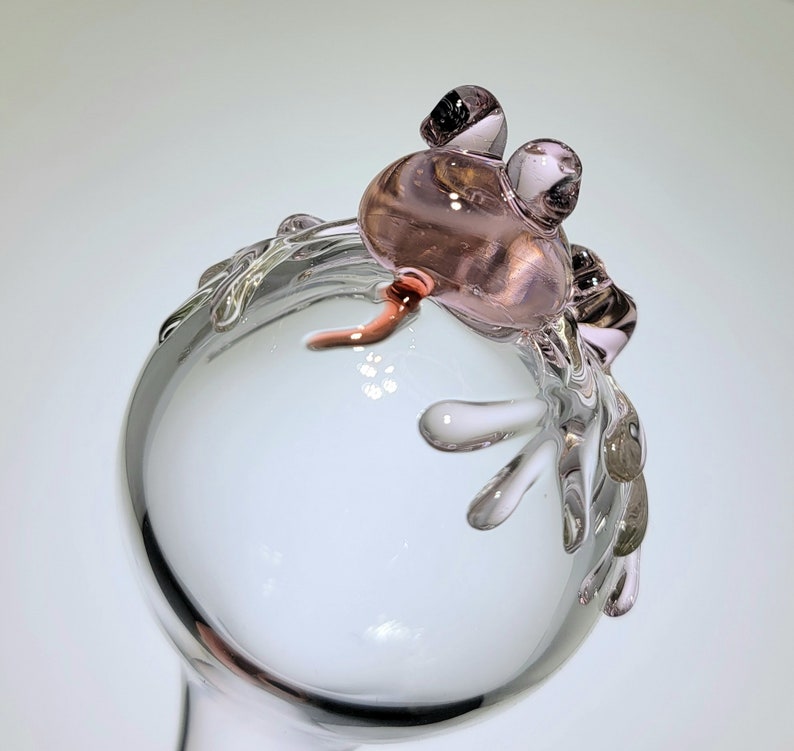 May include: A clear glass sphere featuring a sculpted, light pink glass frog perched on the top edge. The frog has two prominent, rounded eyes and a small, curved tail extending downward. The glass is smooth, transparent, and reflective.