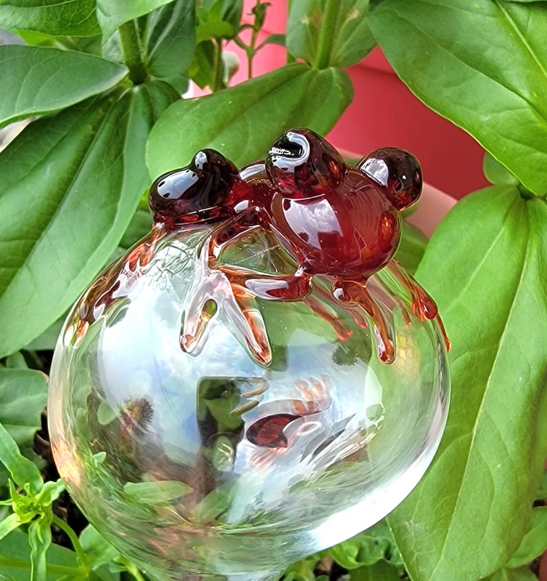 May include: A clear glass watering globe featuring a decorative dark red glass frog perched on top. The frog has a smooth, glossy finish and is fused to the spherical reservoir, which is designed to hold water for plant hydration.