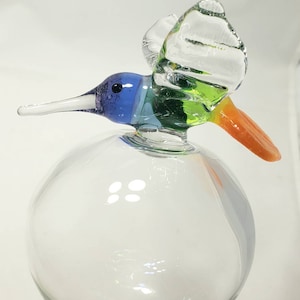 Puede incluir: Un adorno de colibrí de vidrio con un cuerpo transparente, una cabeza azul, alas verdes y una cola naranja. El colibrí está posado en la parte superior de una esfera de vidrio transparente.