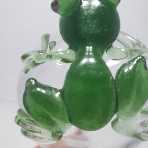 May include: A small, translucent green glass frog figurine with a glittery finish, perched on top of a clear, spherical glass orb. The frog has distinct eyes, four legs with visible toes, and a smooth, rounded body shape crafted from glass.