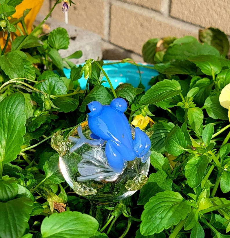 May include: A bright blue glass frog figurine with black eyes, perched atop a clear, spherical glass base that features small, textured, light green glass accents along its bottom edge, creating a decorative garden ornament.