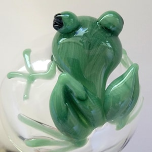 May include: A clear glass stemware piece featuring a sculpted green glass frog perched on the side. The frog has a marbled green finish with dark accents on the eyes and is integrated into the bowl of the glass, showing detailed limbs.