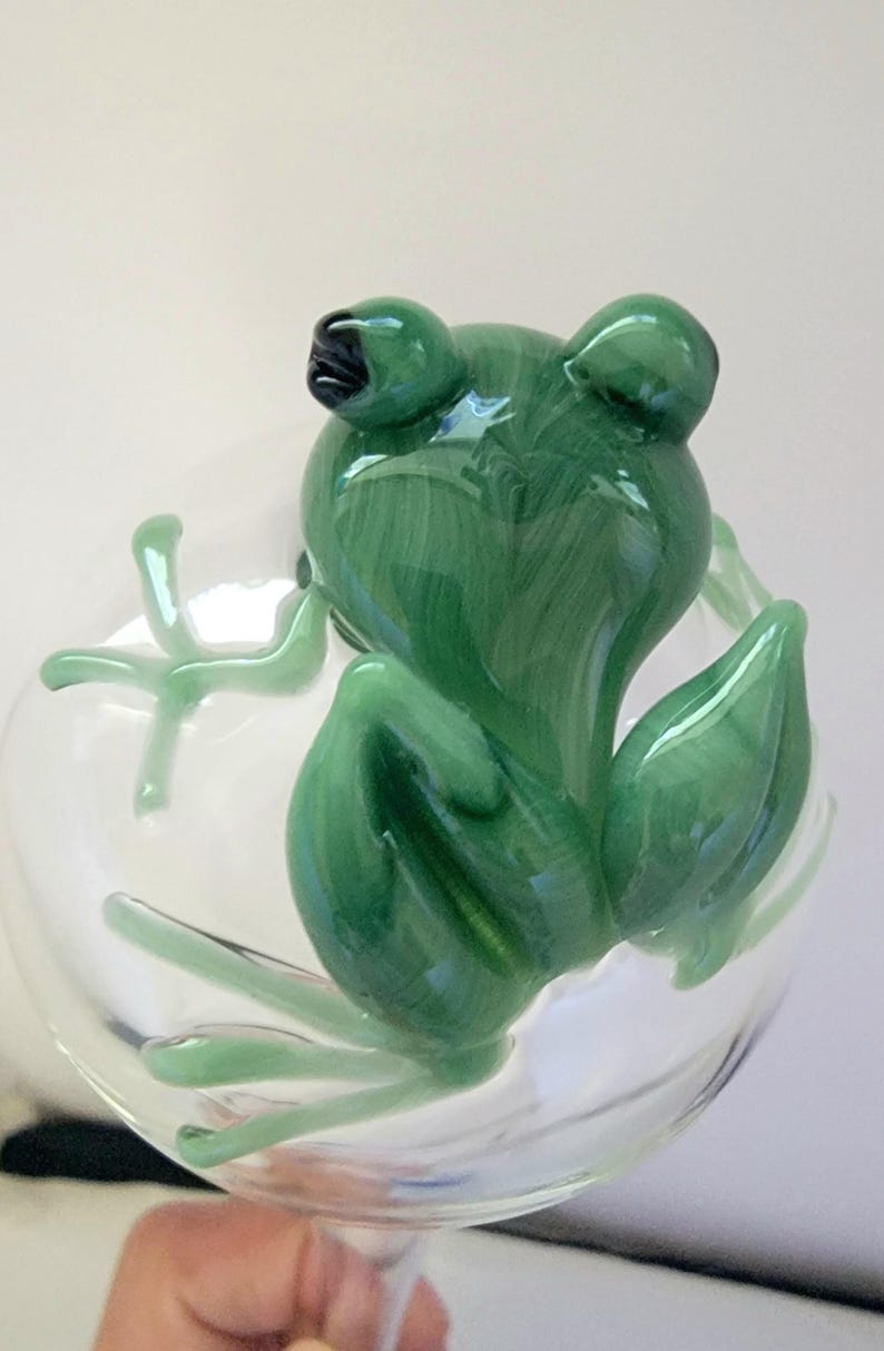 May include: A clear glass stemware piece featuring a sculpted green glass frog perched on the side. The frog has a marbled green finish with dark accents on the eyes and is integrated into the bowl of the glass, showing detailed limbs.