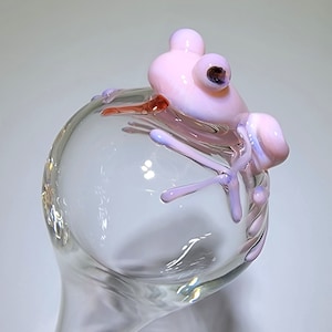 May include: A clear glass sphere topped with a sculpted light pink glass frog. The frog features smooth, rounded limbs and dark, circular eyes, resting its body and legs over the curved surface of the transparent glass orb structure.